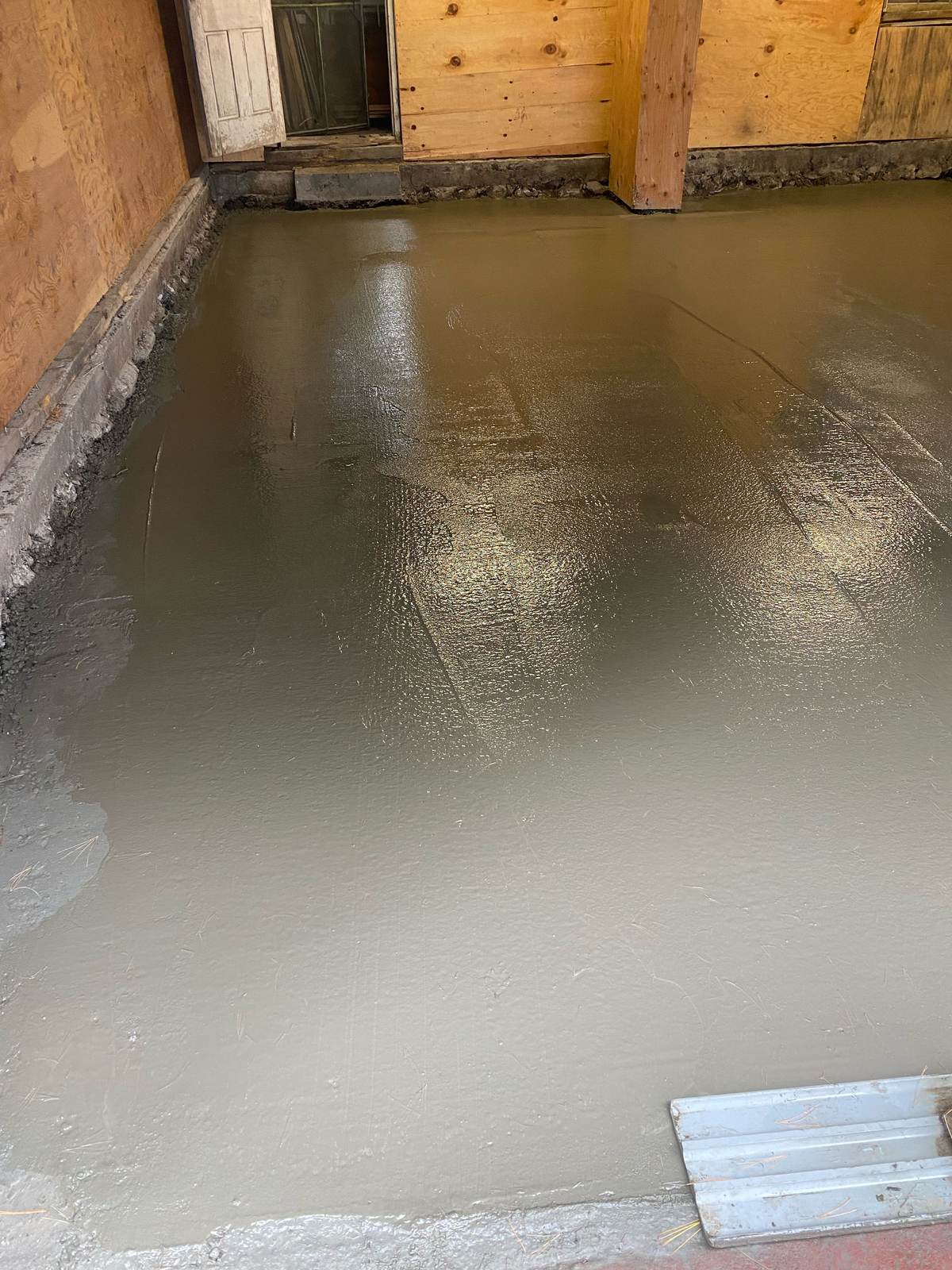Freshly poured and smoothed wet concrete slab inside a garage with plywood walls and a screed tool in the corner
