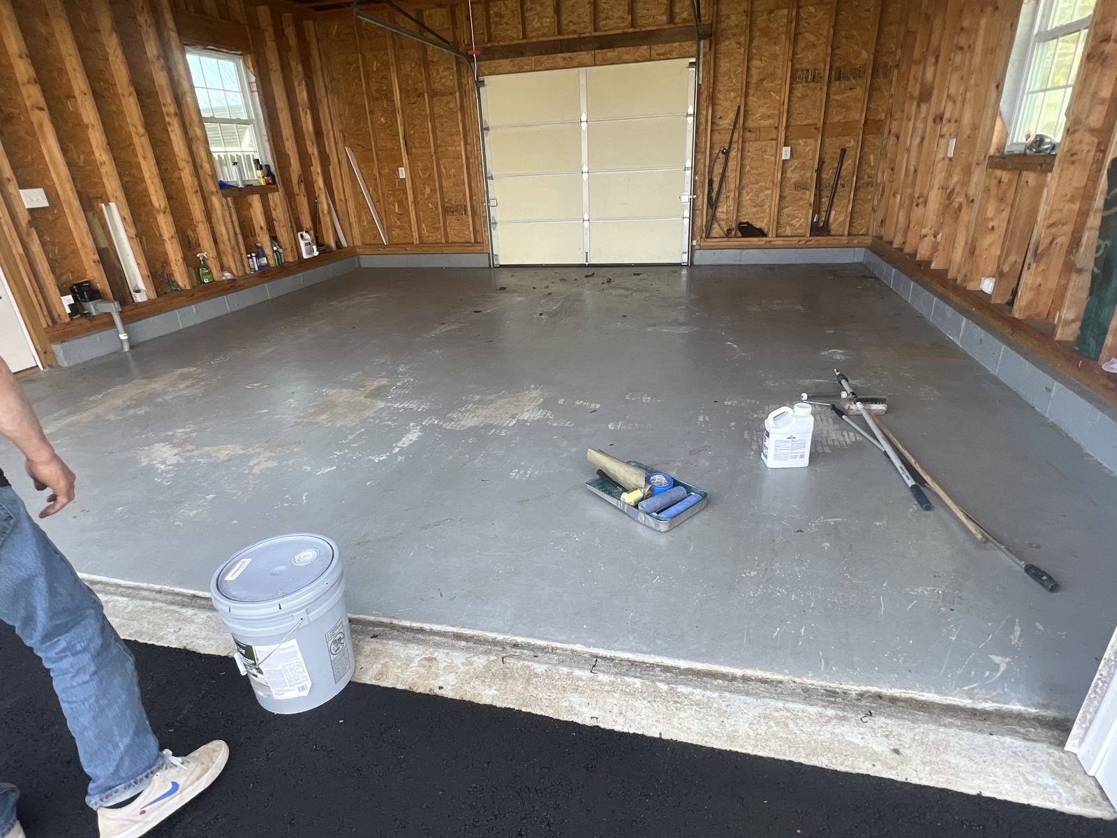 Empty garage with a grey painted concrete floor, a paint bucket, roller tray and applicator tools laid out on the floor