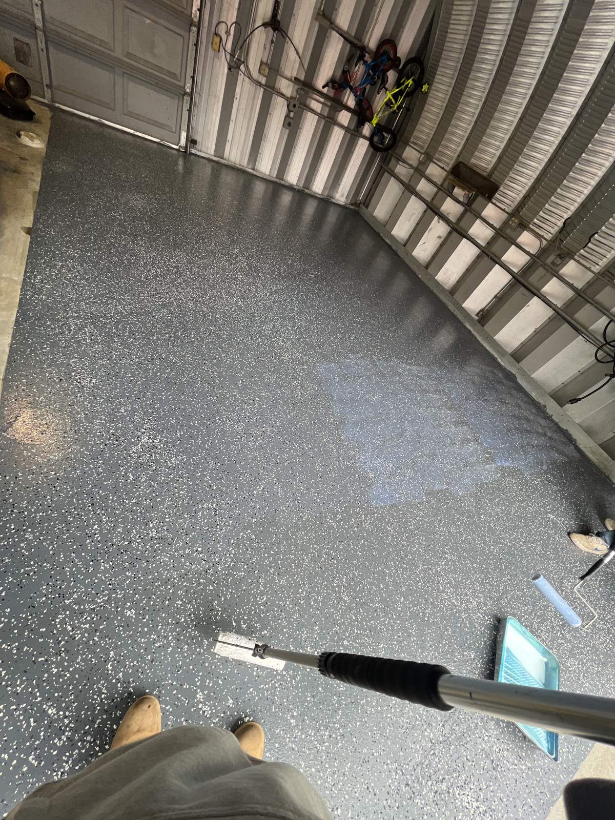 Garage floor with grey flake epoxy and a clear top coat being applied with a long-handled applicator, a wet patch showing a glossy sheen
